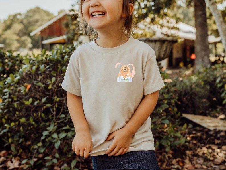 May include: A child wearing a light beige t-shirt with a cartoon bunny wearing glasses and a blue and white striped shirt printed on the front. The child is smiling and standing in front of a green bush.