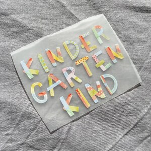 Peut inclure: Une feuille transparente avec les mots "KINDERGARTEN KIND" en lettres color&eacute;es et &agrave; motifs. Les lettres sont de diff&eacute;rentes couleurs, notamment jaune, orange, rose, bleu et vert.