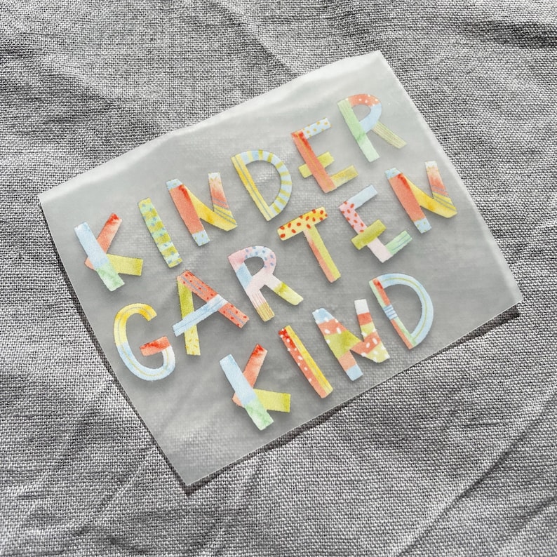 Peut inclure: Une feuille transparente avec les mots "KINDERGARTEN KIND" en lettres color&eacute;es et &agrave; motifs. Les lettres sont de diff&eacute;rentes couleurs, notamment jaune, orange, rose, bleu et vert.