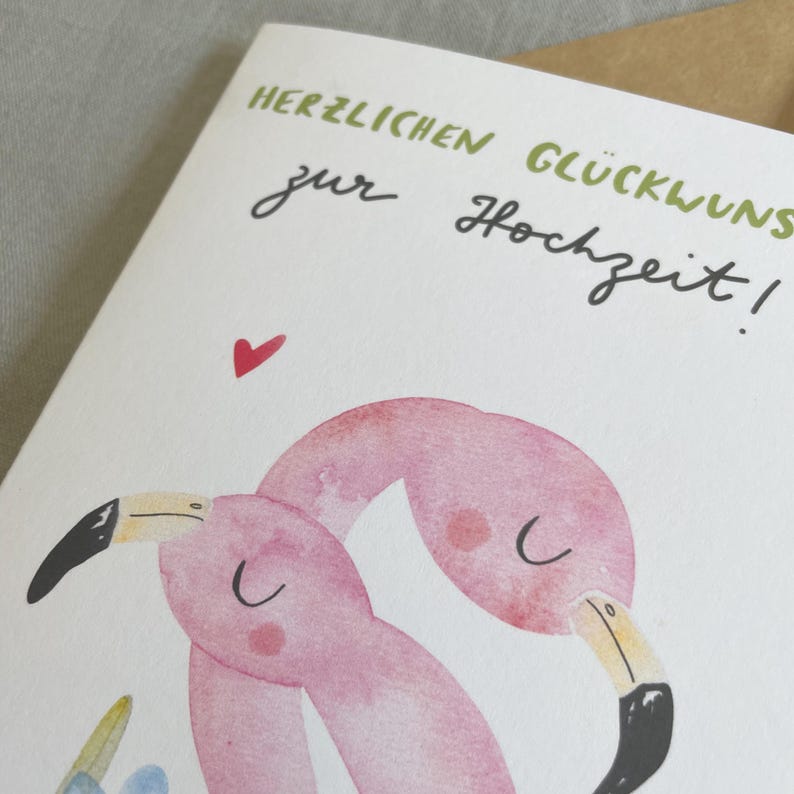 May include: A greeting card featuring a watercolor illustration of two pink flamingos with black beaks and a red heart. The card has the German text "HERZLICHEN GL&Uuml;CKWUNSCH zur Hochzeit!"