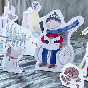 May include: A set of paper Christmas characters, including a person in a wheelchair singing carols, a dog, a tree, and a person wearing a yellow coat. The characters are cut out and ready to be assembled.