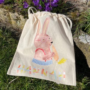 May include: A beige drawstring bag with a cartoon bunny wearing glasses and holding a carrot. The bag has the name "HANNAH" printed on the bottom in colorful letters.
