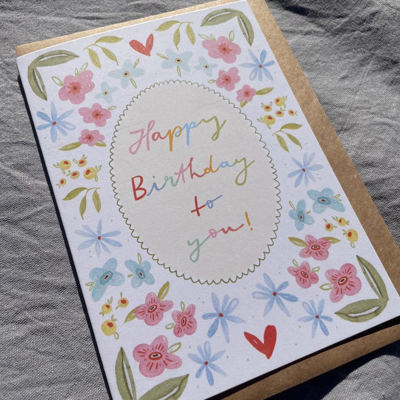 Może przedstawiać: Kartka urodzinowa z kwiatowym wzorem. Karta zawiera tekst "Happy Birthday to you!" w kolorowym skrypcie wewnątrz kropkowatego owalu. Jest ozdobiona r&oacute;żowymi, niebieskimi i ż&oacute;łtymi kwiatami oraz zielonymi liśćmi, z brązową kopertą.