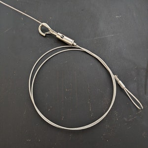 May include: A silver cable with a hook on one end and a loop on the other end. The cable is coiled up on a black surface.