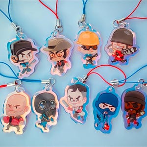 May include: Nine colorful character charms with red and blue strings. The charms are made of clear plastic with a holographic finish and feature characters from the video game Team Fortress 2.