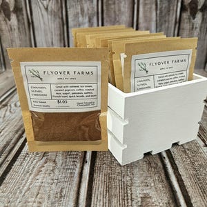 May include: A white wooden crate filled with small brown paper bags containing a spice blend labeled "FLYOVER FARMS Apple Pie Spice". The label lists the ingredients as cinnamon, nutmeg, and cardamom. The price is $1.05.