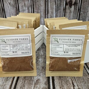 May include: Two small brown paper bags filled with spices. The bags are labeled "Flyover Farms" and list the ingredients and price. The bag on the left is labeled "Apple Pie Spice" and the bag on the right is labeled "Pumpkin Pie Spice".