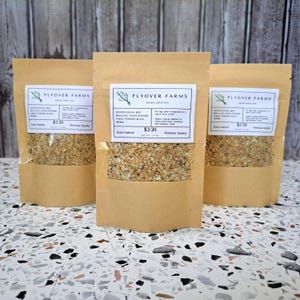 May include: Three brown paper bags with white labels that say "Flyover Farms Onion Soup Mix". The labels also include the ingredients, "Minced Onion, Beef Bouillon, Onion Powder, Garlic Powder, Black Pepper", and the price, "$3.20". The bags are filled with a light brown, granular mix.