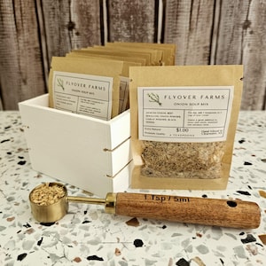 May include: A brown paper packet of Flyover Farms Onion Soup Mix with a wooden scoop filled with dried onion flakes. The packet lists the ingredients: minced onion, beef bouillon, onion powder, garlic powder, black pepper. The packet also states that it makes 4 teaspoons and costs $1.00.