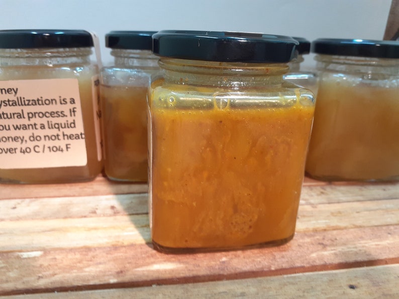 May include: Four glass jars filled with honey. The jar in the center is filled with a darker, more viscous honey. The label on the jar on the left reads "Honey Crystallization is a natural process. If you want a liquid honey, do not heat over 40 C / 104 F."