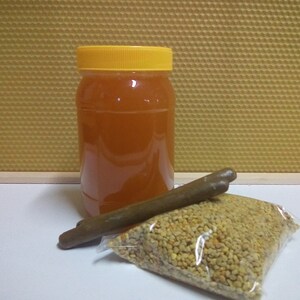 May include: A clear glass jar with a yellow lid filled with honey, a brown stick, and a bag of yellow bee pollen.