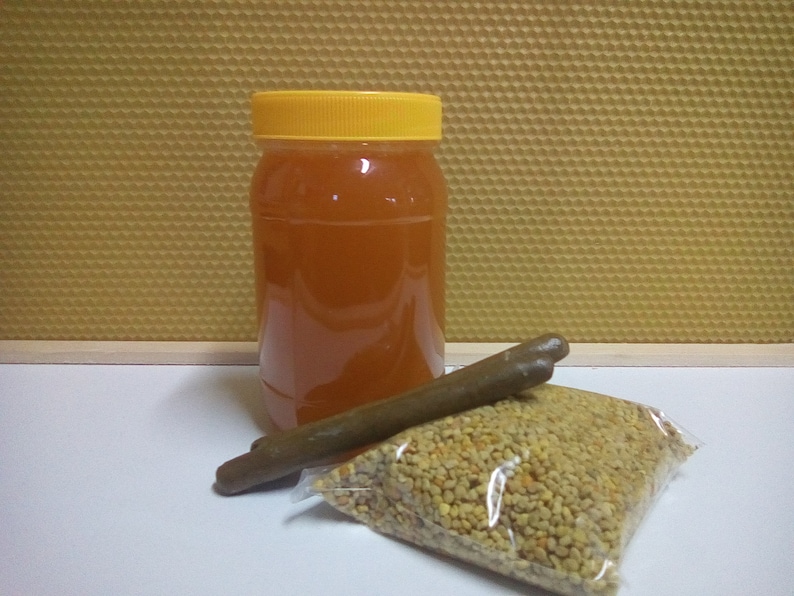 May include: A clear glass jar with a yellow lid filled with honey, a brown stick, and a bag of yellow bee pollen.