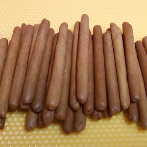 Puede incluir: Un montón de galletas marrones en forma de palitos, dispuestas sobre una superficie amarilla texturizada. Las galletas tienen un color y forma uniformes, con extremos redondeados. La imagen está bien iluminada, resaltando la textura de las galletas.