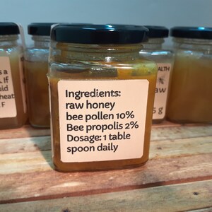May include: A glass jar filled with a golden honey-like substance. The label reads "Ingredients: raw honey bee pollen 10% Bee propolis 2% Dosage: 1 table spoon daily".