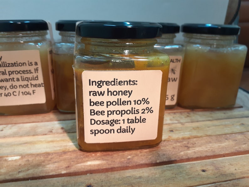 May include: A glass jar filled with a golden honey-like substance. The label reads "Ingredients: raw honey bee pollen 10% Bee propolis 2% Dosage: 1 table spoon daily".