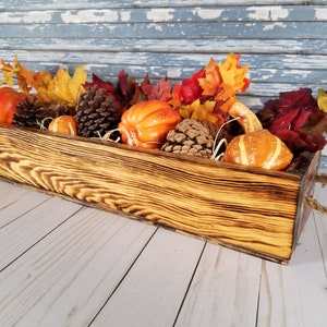 Könnte beinhalten: Eine rustikale Holzkiste mit einem Seilgriff, gefüllt mit künstlichen Kürbissen, Tannenzapfen und Herbstblättern. Die Kiste ist dunkelbraun gebeizt und hat einen Used-Look.