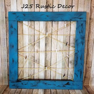 Puede incluir: Un marco de madera azul con una cuadrícula de cuerda y pinzas para la ropa. El marco está desgastado y tiene un aspecto rústico. El texto "J25 Rustic Decor" es visible en la parte superior del marco.