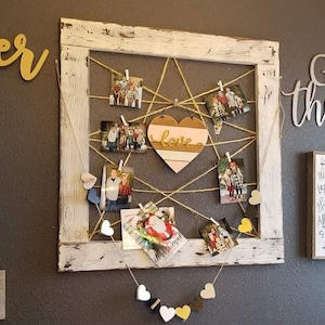 May include: A whitewashed wooden frame with a string grid holding photos and a pink heart with the word "love" on it. A string of wooden hearts in various colors hangs below the frame.