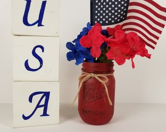 以下が含まれることがあります： 赤いメイソンジャーに、青と赤の花の飾り付けと小さなアメリカ国旗が飾られています。ジャーは紐で飾られており、「Ball Mason」の文字が見えます。背景には、「U」、「S」、「A」の文字が青で書かれた白いブロックが3つあります。