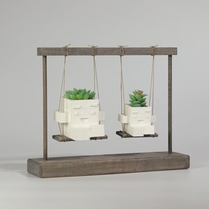 May include: Two white ceramic planters shaped like faces with smiling expressions are hanging from a wooden swing set. Each planter has a small green succulent plant growing inside.
