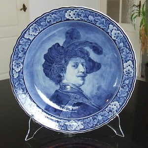 May include: A blue and white ceramic plate with a portrait of a man in a feathered hat. The plate has a decorative border with floral and leaf designs.