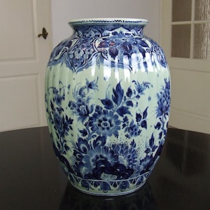 Puede incluir: Un jarrón de cerámica grande con un cuerpo ancho y un cuello estrecho. El jarrón presenta un fondo verde claro con intrincados diseños florales azules. El borde tiene un borde festoneado. Una pieza decorativa para el hogar.