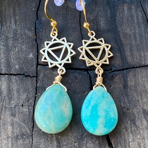 Puede incluir: Pendientes de oro con una piedra verde turquesa y un sol de oro con un triángulo en su interior. Los pendientes están sobre un fondo de madera oscura.