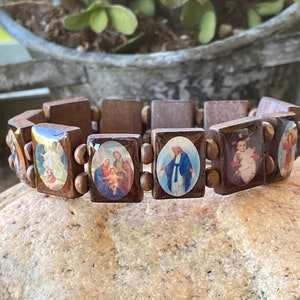 May include: A brown wooden bracelet with oval-shaped images of religious figures. The images include a family, a woman in blue, a baby, and an angel.