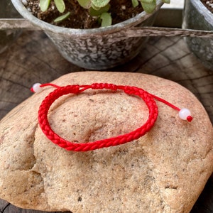 May include: A red string bracelet with white beads on a brown rock. The bracelet is made of a braided cord.