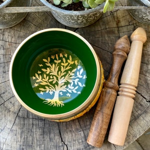 May include: A green singing bowl with a gold tree design and two wooden strikers. The bowl is sitting on a wooden surface.
