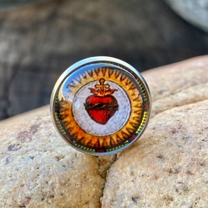 Könnte beinhalten: Eine silberfarbene Anstecknadel mit einem runden Glascabochon, der ein Vintage-Bild eines roten Herzens mit Dornenkrone und Kreuz zeigt, umgeben von einem goldenen Sonnenstrahlen-Design.