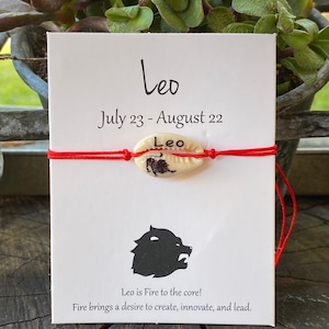 May include: A white card with red string and a shell charm with the zodiac sign Leo. The card reads "Leo, July 23 - August 22, Leo is Fire to the core! Fire brings a desire to create, innovate, and lead."