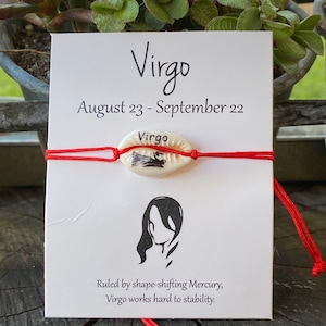 May include: A white card with the word "Virgo" and the dates August 23 - September 22. A red string bracelet with a cowrie shell charm is in front of the card. The charm has the word "Virgo" and a black illustration.