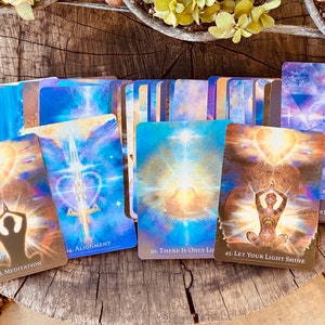 May include: A set of colorful oracle cards with images of people meditating, a sword, and a heart. The cards have numbers and titles, such as "8. Meditation", "14. Alignment", "30. There is Only Li", and "45. Let Your Light Shine".