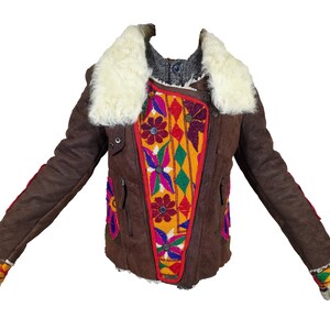 K&ouml;nnte beinhalten: Braune Jacke mit wei&szlig;em Pelzkragen und farbenfrohen Stickereidetails. Die Jacke hat ein einzigartiges Design mit leuchtenden Blumen- und geometrischen Mustern auf der Vorderseite und den &Auml;rmeln. Die &Auml;rmel haben rote Verzierungen und die Manschetten sind mit wei&szlig;em Fell besetzt.