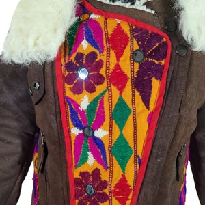 K&ouml;nnte beinhalten: Braune Jacke mit cremefarbenem Fellkragen und farbenfrohen Stickereidetails. Die Jacke hat ein Steppmuster an den &Auml;rmeln und ein lebendiges, geometrisches Design auf der Vorderseite mit roten, gelben, lila und gr&uuml;nen Akzenten. Sie hat schwarze Kn&ouml;pfe und Rei&szlig;verschl&uuml;sse.