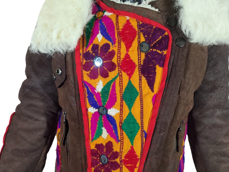 K&ouml;nnte beinhalten: Braune Jacke mit cremefarbenem Fellkragen und farbenfrohen Stickereidetails. Die Jacke hat ein Steppmuster an den &Auml;rmeln und ein lebendiges, geometrisches Design auf der Vorderseite mit roten, gelben, lila und gr&uuml;nen Akzenten. Sie hat schwarze Kn&ouml;pfe und Rei&szlig;verschl&uuml;sse.