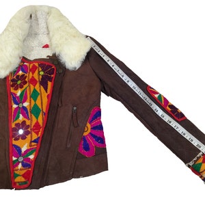 K&ouml;nnte beinhalten: Braune Wildlederjacke mit cremefarbenem Shearling-Kragen und -Futter. Die Jacke hat bunte, gestickte Blumen- und geometrische Designs auf der Vorderseite und den &Auml;rmeln. Die &Auml;rmell&auml;nge betr&auml;gt ca. 61 cm.