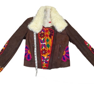 K&ouml;nnte beinhalten: Braune Wildlederjacke mit cremefarbenem Kunstfellkragen. Die Jacke hat bunte, gestickte Blumen- und geometrische Muster auf der Vorderseite, den &Auml;rmeln und den Manschetten. Ein Ma&szlig;band ist auf der Jacke platziert. Die Jacke hat einen Rei&szlig;verschluss.