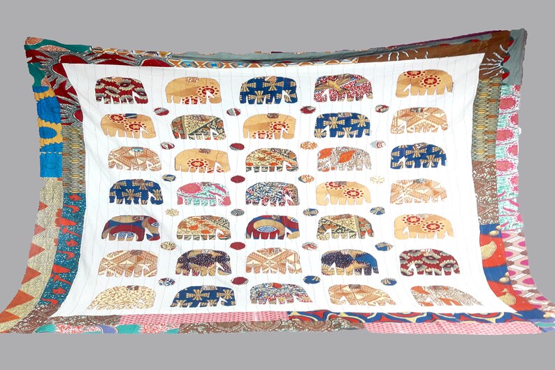 K&ouml;nnte beinhalten: Wei&szlig;e Baumwolldecke mit Patchwork-Bord&uuml;re. Die Decke zeigt ein sich wiederholendes Muster aus wei&szlig;en Elefanten mit farbenfrohen gemusterten K&ouml;rpern. Die Elefanten sind in Reihen angeordnet und von kleinen, bunten Kreisen umgeben.