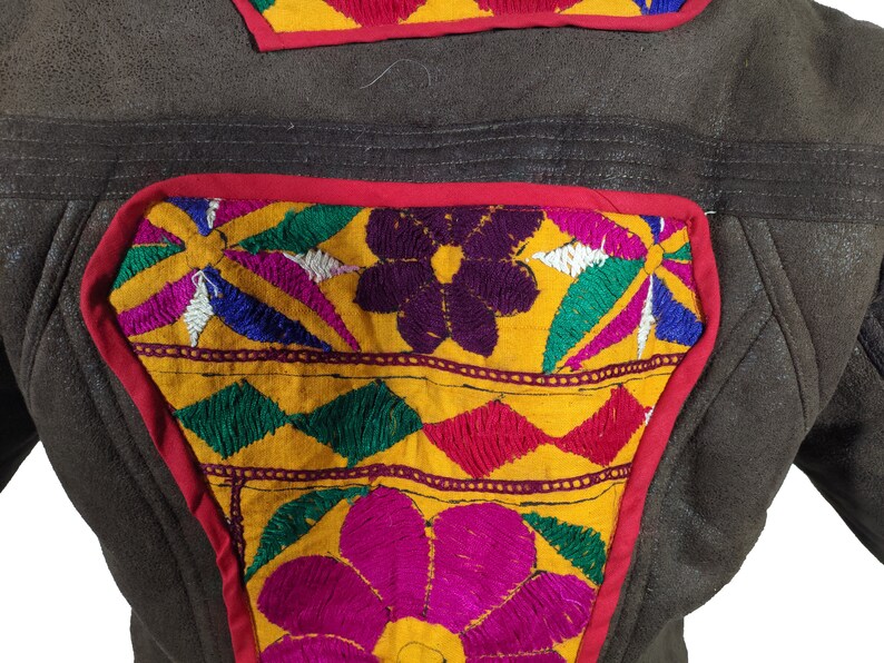 K&ouml;nnte beinhalten: Nahaufnahme einer braunen Lederjacke mit bunten gestickten Aufn&auml;hern. Die Aufn&auml;her zeigen florale und geometrische Designs in Gelb-, Rosa-, Gr&uuml;n-, Blau- und Lilat&ouml;nen, umrandet in Rot.
