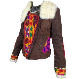 K&ouml;nnte beinhalten: Braune Wildlederjacke mit wei&szlig;em Kunstfellkragen und bunten, gestickten Blumen- und geometrischen Mustern. Die Jacke hat einen diagonalen Rei&szlig;verschluss und dekorative Verzierungen an den &Auml;rmeln.