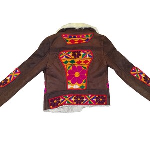 K&ouml;nnte beinhalten: Braune Wildlederjacke mit farbenfrohen, gestickten Blumen- und geometrischen Designs. Die Jacke hat einen cremefarbenen Kunstfellkragen und -futter. Die &Auml;rmel und der R&uuml;cken sind mit leuchtenden Mustern in Rot, Gelb, Blau und Lila verziert.