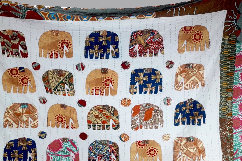 K&ouml;nnte beinhalten: Eine wei&szlig;e Baumwolldecke mit Patchwork-Muster, die mehrere braune, blaue und rote Elefanten zeigt. Die Elefanten sind aus verschiedenen gemusterten Stoffen gefertigt und in einem Gittermuster angeordnet.