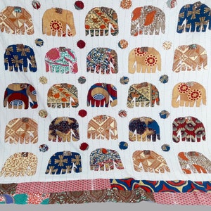 K&ouml;nnte beinhalten: Wei&szlig;e Baumwolldecke mit Patchwork-Bord&uuml;re. Die Decke zeigt ein sich wiederholendes Muster aus bunten Elefanten-Silhouetten.