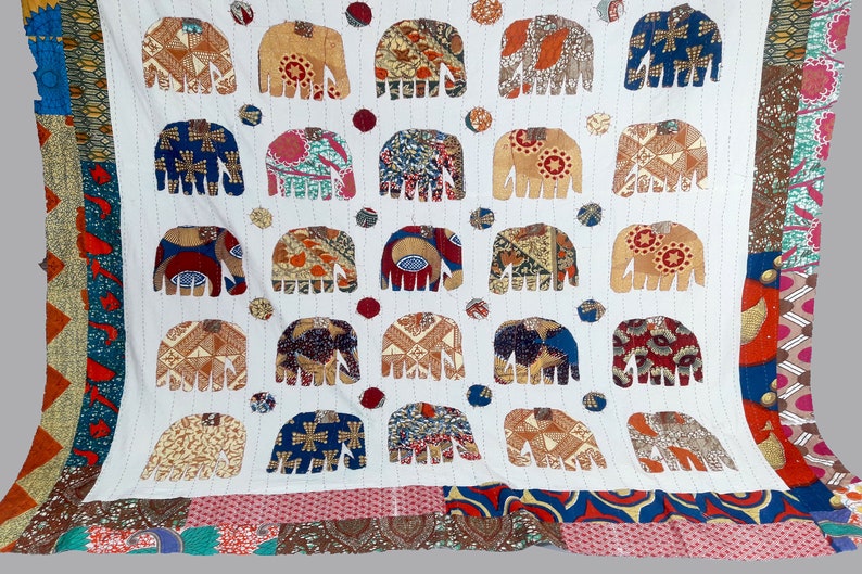 K&ouml;nnte beinhalten: Wei&szlig;e Baumwolldecke mit Patchwork-Bord&uuml;re. Die Decke zeigt ein sich wiederholendes Muster aus bunten Elefanten-Silhouetten.