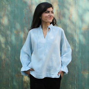 May include: A light blue linen blouse with long sleeves and a V-neck. The blouse features a ruffled cuff and a tie at the neckline. The model is wearing black pants.