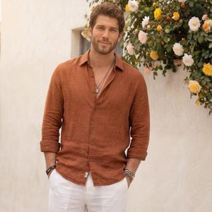 Puede incluir: Un hombre con una camisa abotonada de color óxido y pantalones blancos. La camisa tiene mangas largas y está desabrochada en la parte superior. Lleva un collar y pulseras. El fondo presenta una pared blanca y flores.