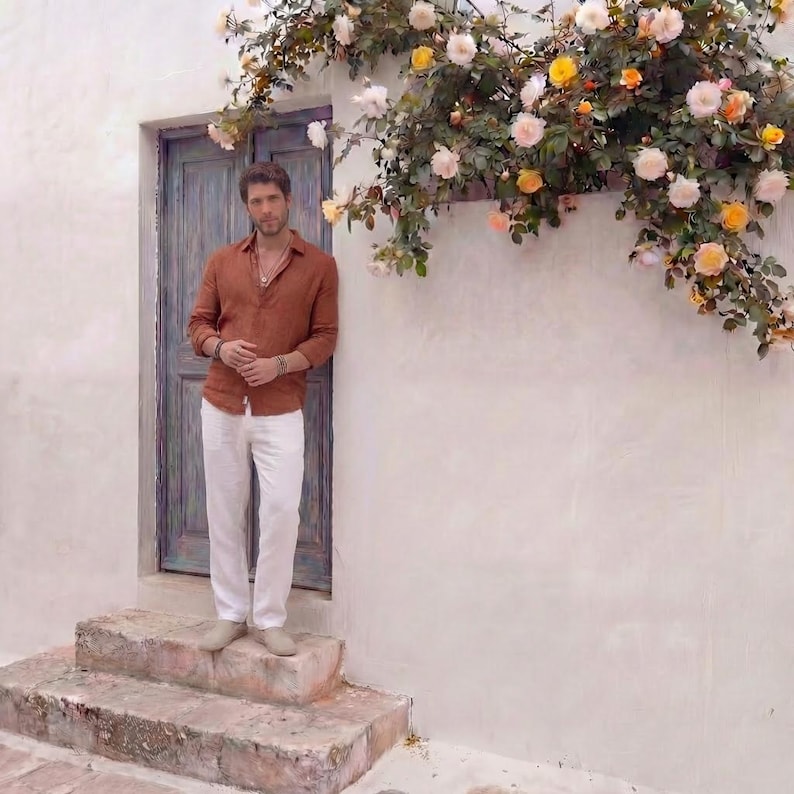 May include: A person wearing a rust-colored button-down shirt and white pants stands on stone steps next to a weathered blue door. The wall is white, and a floral arrangement of roses in shades of pink, yellow, and orange cascades above.