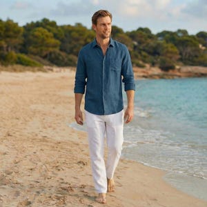 May include: A man wearing a dark blue linen shirt and white linen pants walks on a sandy beach. The shirt has a single chest pocket. The background features a shoreline, trees, and a calm sea.